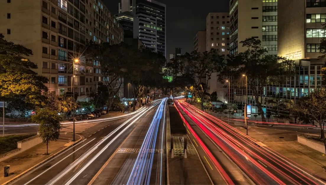 Avenida movimentada à noite com rastros de luz dos veículos entre prédios